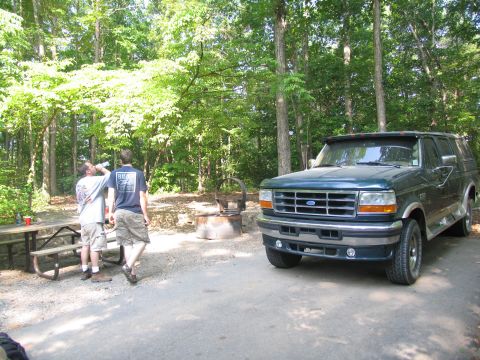 1995 Ford C-350 (Centurion Conversions) - Two people stand by a picnic table near a green Ford Bronco in a wooded campground.