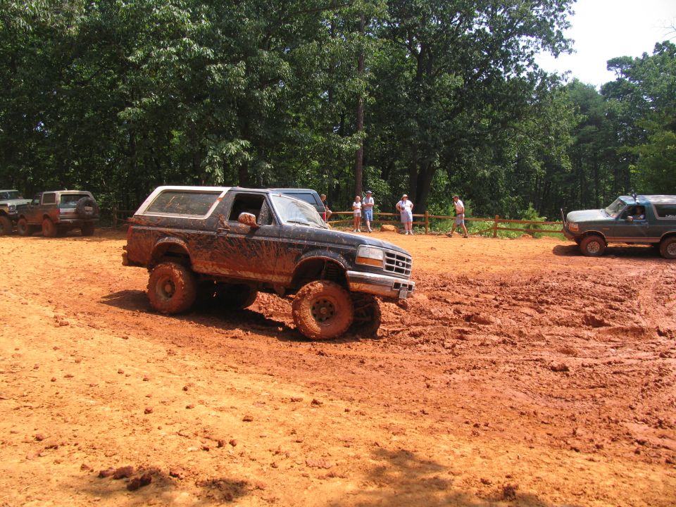 Muddy Ford Bronco driving through a dirt track with other vehicles and people in the background