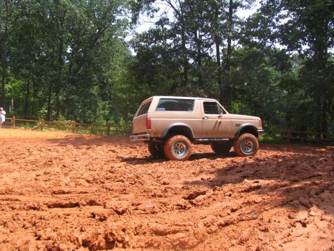 1995 Ford C-350 (Centurion Conversions) - A tan Ford Bronco navigates a muddy terrain, its lifted suspension and off-road tires clearly visible.
