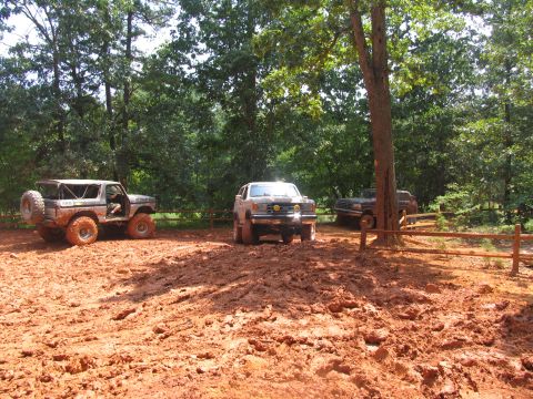 1995 Ford C-350 (Centurion Conversions) - Multiple off-road vehicles are parked in a muddy clearing surrounded by trees.