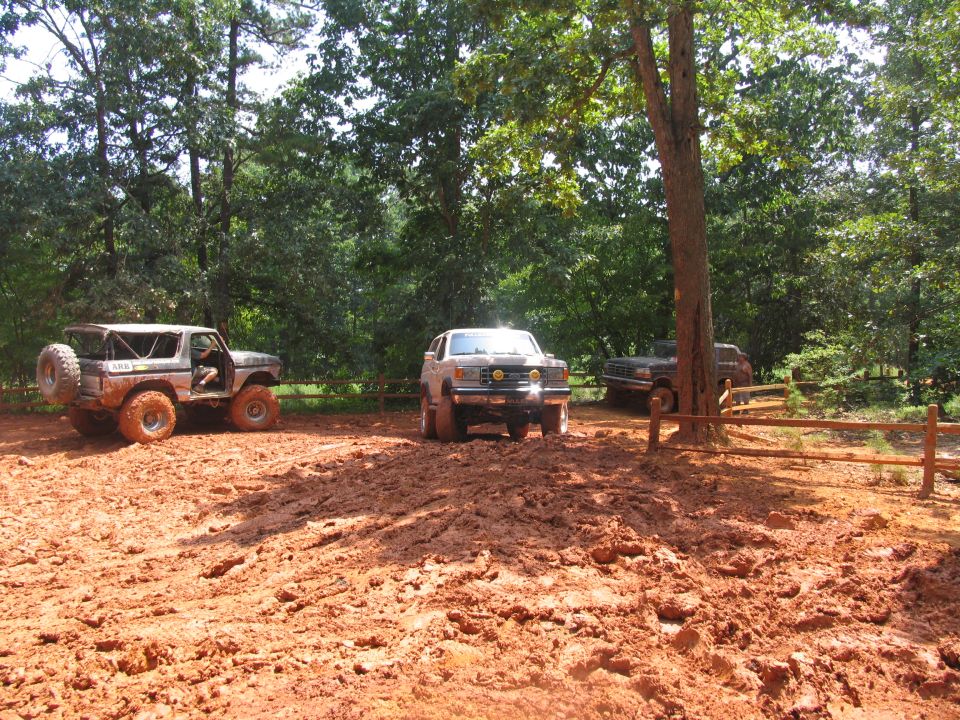 Three off-road vehicles parked in a muddy area with trees and a wooden fence