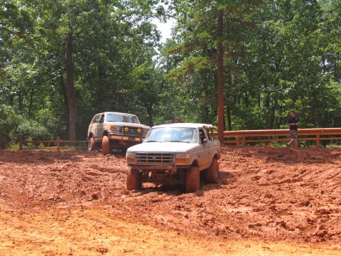 1995 Ford C-350 (Centurion Conversions) - Two SUVs navigate a muddy off-road course with trees and a wooden fence in the background.