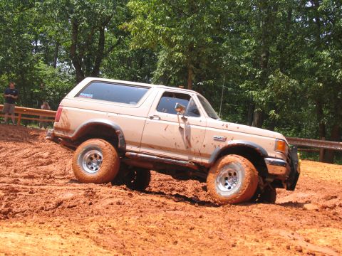 1995 Ford C-350 (Centurion Conversions) - A tan Ford Bronco is shown struggling through a muddy off-road trail, with trees and spectators in the background.