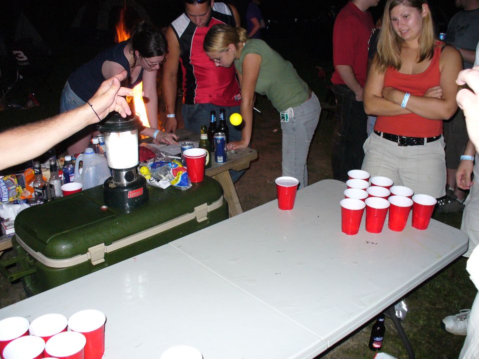 people gathered around tables with red cups set up for a game, some holding drinks, with a lantern and cooler nearby