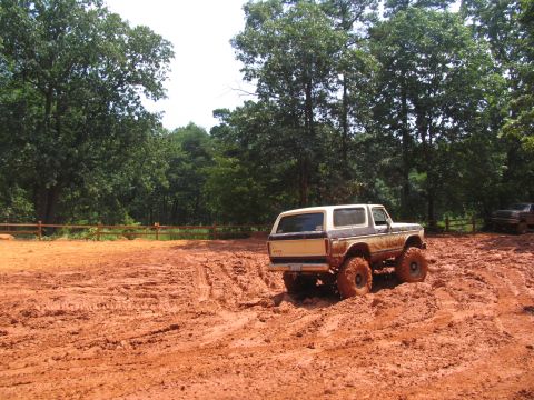 1995 Ford C-350 (Centurion Conversions) - A lifted Ford Bronco navigates a muddy off-road course with trees and a wooden fence in the background.