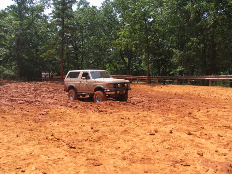 1995 Ford C-350 (Centurion Conversions) - A tan Ford Bronco navigates a muddy terrain with trees in the background.