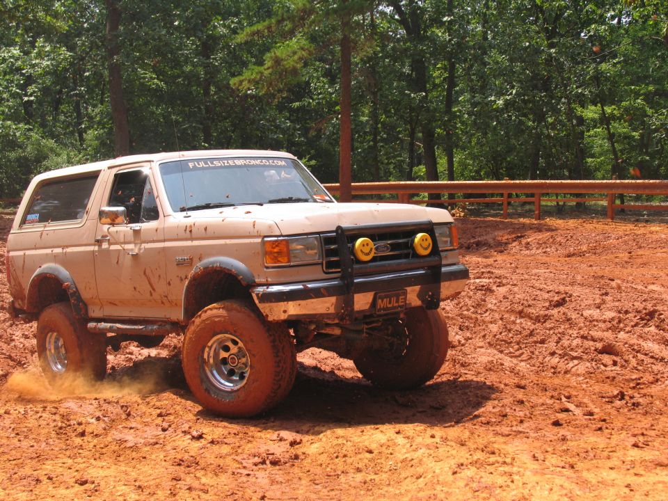 Tan Ford Bronco with off-road tires driving through mud, front bull bar with smiley face lights