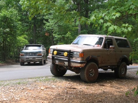 1995 Ford C-350 (Centurion Conversions) - Two trucks, a tan Ford Bronco and a green Ford F-Series, are parked on a dirt road surrounded by trees.