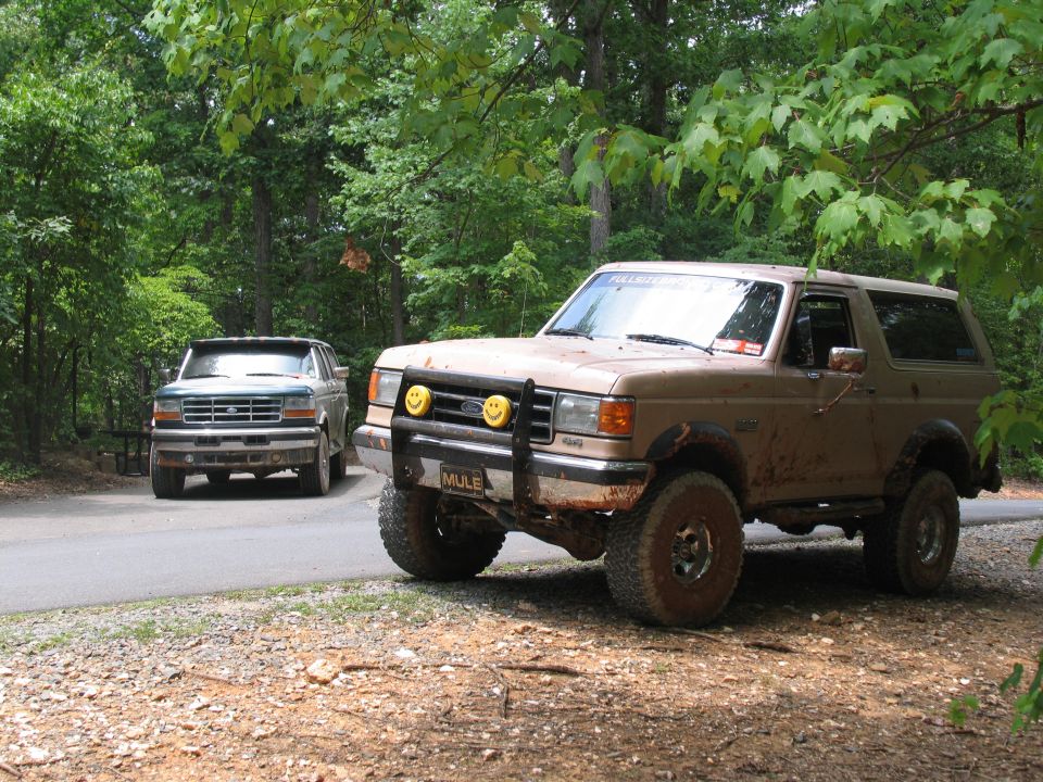 Tan Ford Bronco with off-road tires and bumper, mud-splattered, parked on dirt road. Another truck is visible behind it