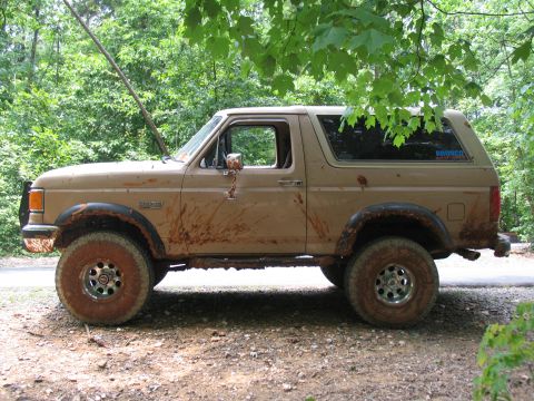 1995 Ford C-350 (Centurion Conversions) - A mud-covered tan Ford Bronco is seen from the side, parked on a dirt road under trees.