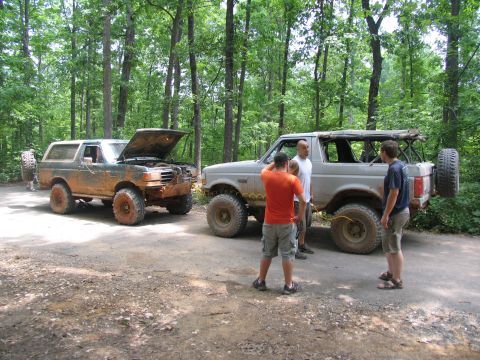 1995 Ford C-350 (Centurion Conversions) - Two off-road modified Ford Broncos are parked on a dirt path surrounded by trees, with people gathered near them.