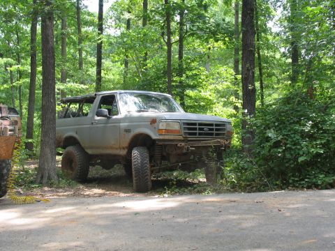 1995 Ford C-350 (Centurion Conversions) - A lifted Ford Bronco sits off-road in a wooded area, its front bumper removed.