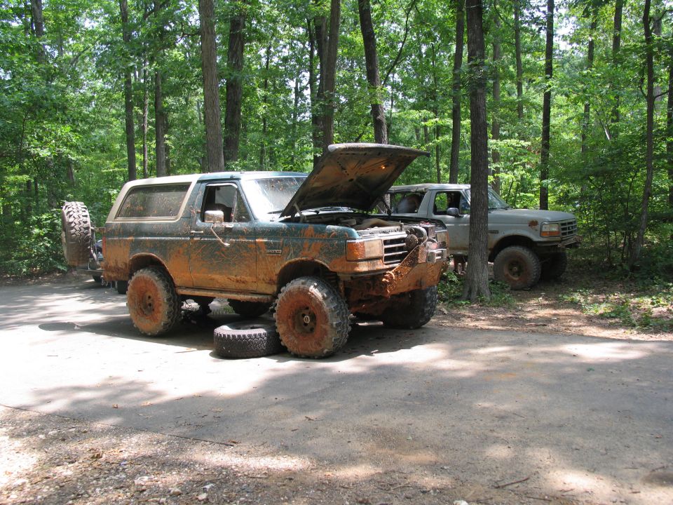 Mud-covered teal Ford Bronco with hood open, spare tire mounted on rear, and a second truck behind it