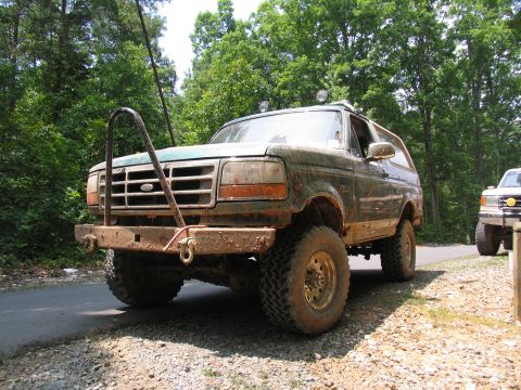 1995 Ford C-350 (Centurion Conversions) - Mud-covered green Ford Bronco parked on a gravel shoulder next to a paved road, with a second truck in the background.