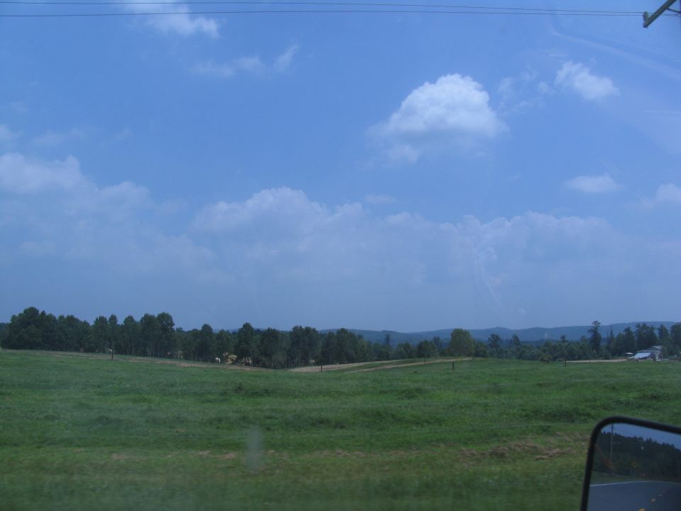 Side mirror view of a rural landscape with green fields and trees under a blue sky