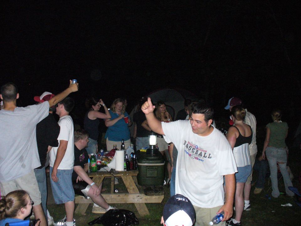 People gathered outdoors at night, some holding drinks and talking near a picnic table