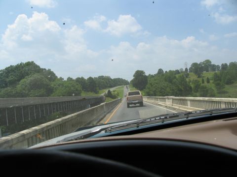 1995 Ford C-350 (Centurion Conversions) - A brown pickup truck travels down a highway on a sunny day, with trees and hills in the background.