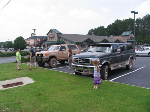 1995 Ford C-350 (Centurion Conversions) - Two Ford Broncos are parked in a lot outside a building with people standing around them.