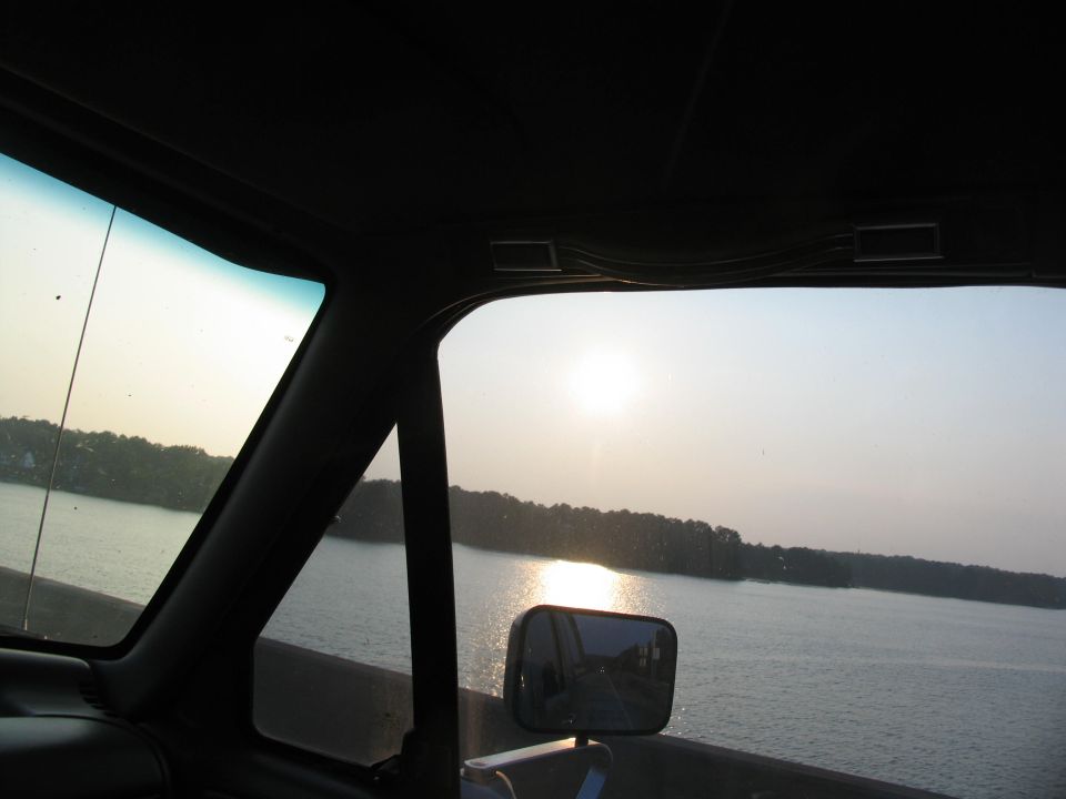 View from inside truck cab, looking out windshield at lake and trees. Sun is low in the sky