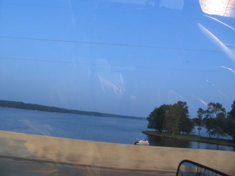 1995 Ford C-350 (Centurion Conversions) - A boat is docked near a small island with trees, viewed from a moving vehicle on a clear day.