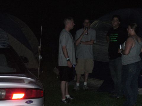 1991 Ford Probe - People gather around a tent and a car at night, with a campsite visible in the background.
