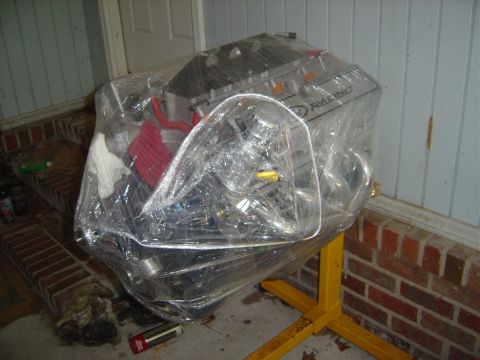 1986 Ford Bronco - Engine wrapped in plastic on a yellow engine stand, with a brick wall and door in the background.