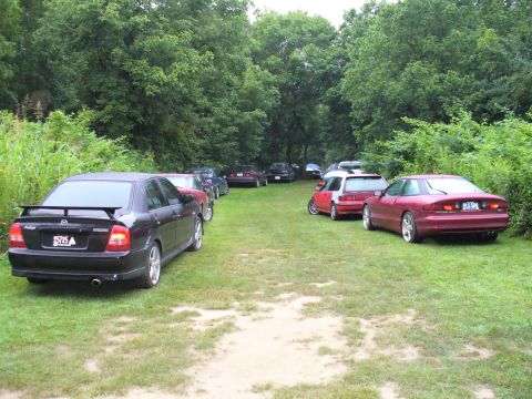 1991 Ford Probe - A line of cars, including a 1991 Ford Probe, are parked on a grassy field surrounded by trees.