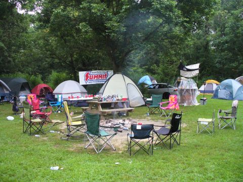 1991 Ford Probe - A busy campsite is set up with multiple tents, chairs arranged around a fire pit, and a picnic table laden with drinks.
