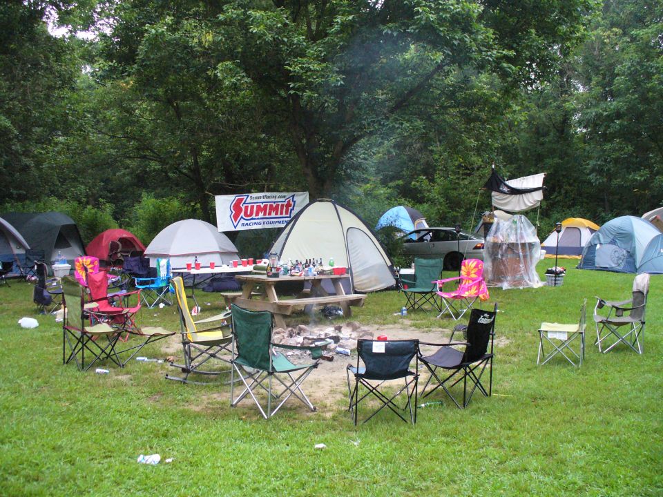 Campsite scene with tents, picnic table, campfire, and chairs. A car is parked in the background