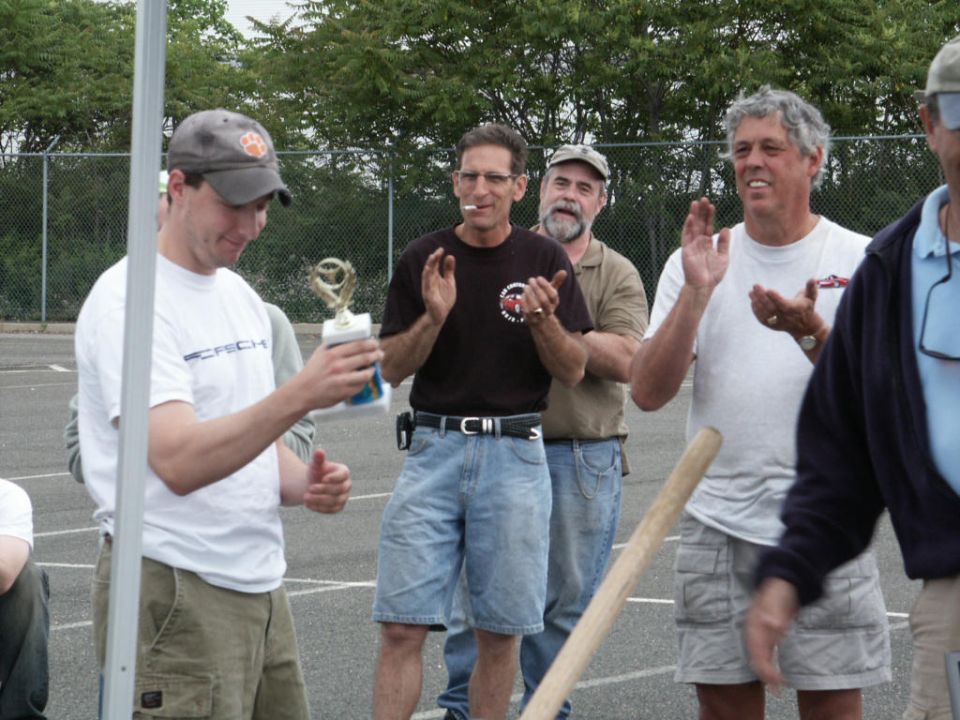 Man in white t-shirt holding trophy, other men clapping. SuperMotors logo visible