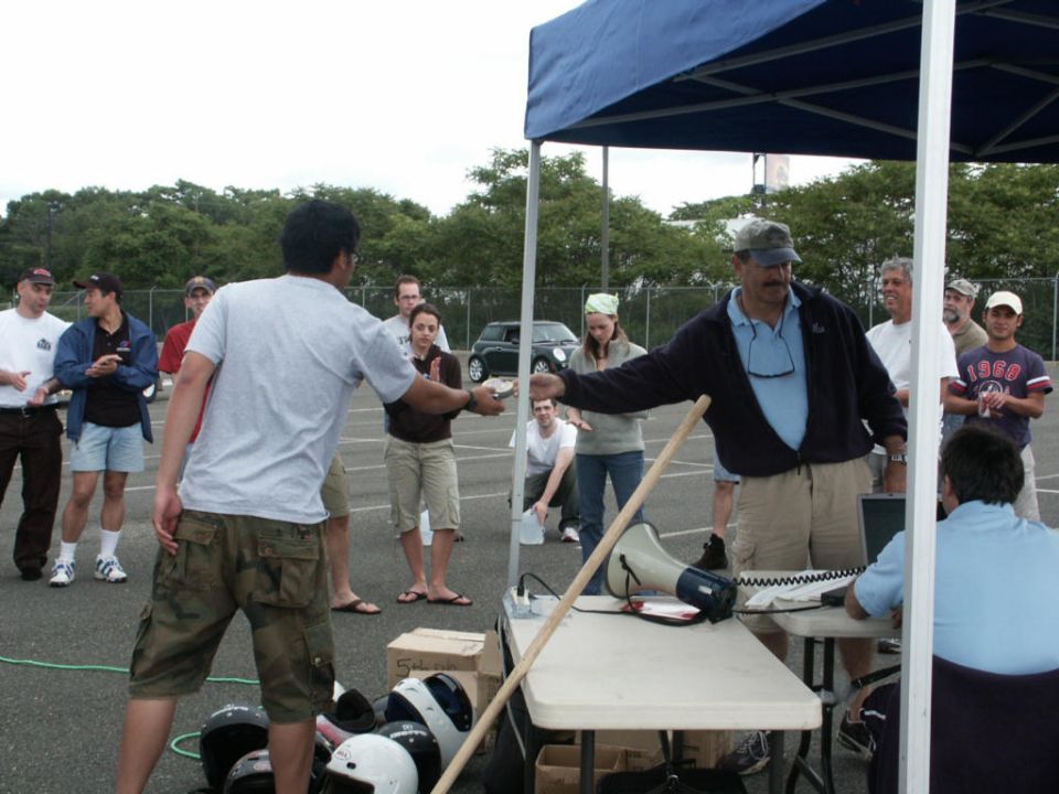 People are gathered around a table with a megaphone and a laptop, possibly at a race or event. A black car is visible in the background