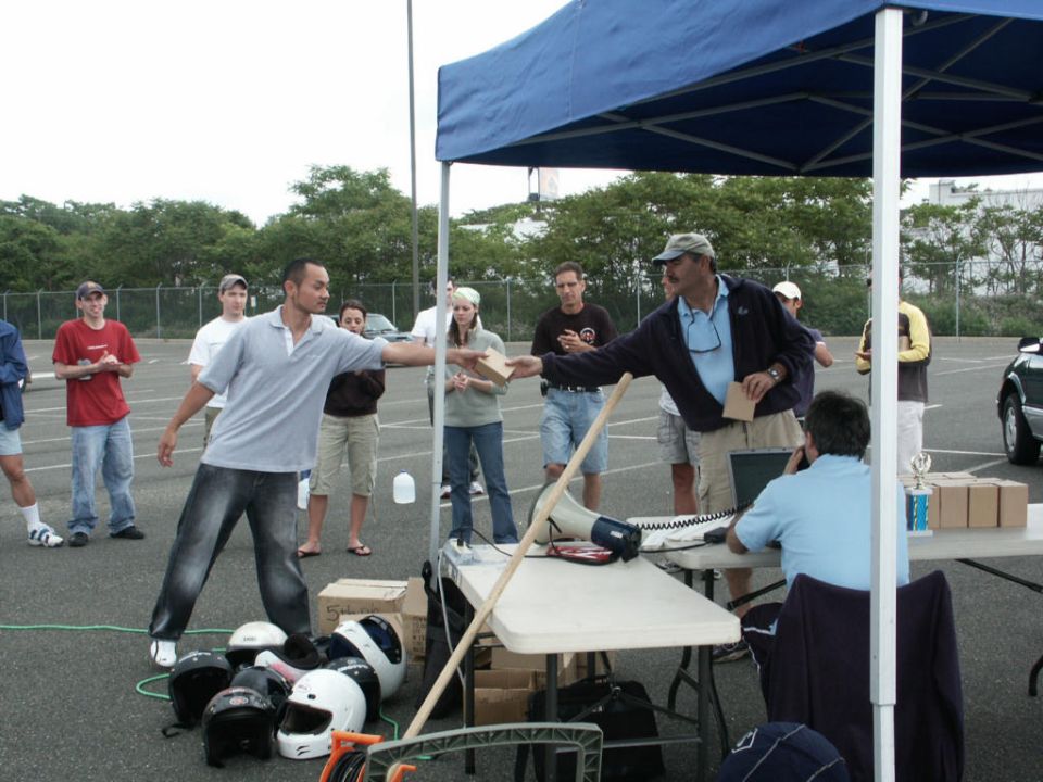 People gathered around tables at an outdoor event, with helmets on the ground and a blue tent overhead