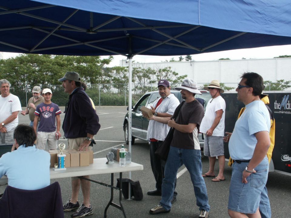 Men gathered under a blue canopy at an outdoor event, with a table displaying trophies and boxes