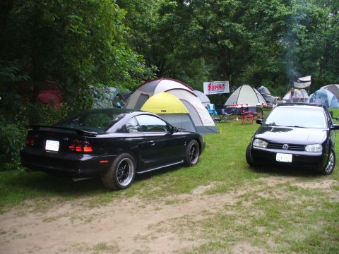 1991 Ford Probe - A black coupe with a prominent spoiler is parked on a dirt path next to a Volkswagen, with tents in the background.