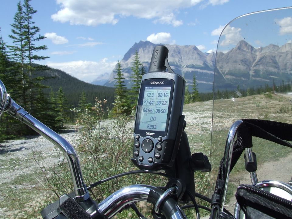Motorcycle handlebars with Garmin GPS unit mounted, showing trip data against a mountain landscape