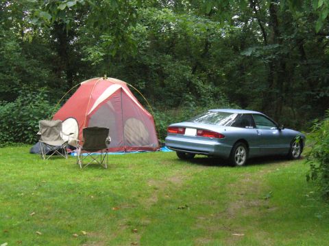 1991 Ford Probe - A blue coupe is parked on the grass next to a red tent and two camping chairs in a wooded area.