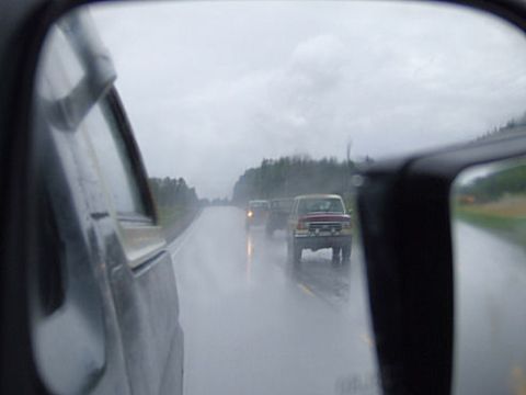 1988 Ford Bronco - Shot forward through a rain-streaked side window and door frame of a light-colored truck, showing a wet two-lane highway with a lifted red-…