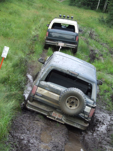 1988 Ford Bronco - Two mud-covered full-size Ford Broncos on a deeply rutted muddy trail cut through a green grassy hillside, viewed from above and behind. Th…