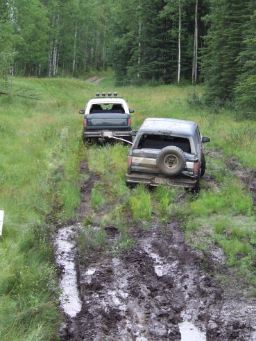 1988 Ford Bronco - Two SUVs navigate a challenging muddy off-road trail surrounded by trees.