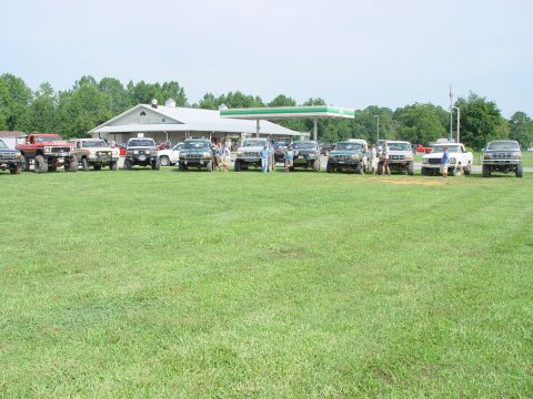 1996 Ford Bronco - Multiple SUVs and trucks are parked in a grassy field near a building and gas station.