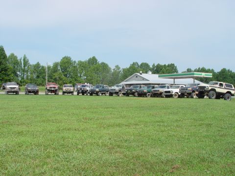 1996 Ford Bronco - Multiple SUVs and trucks are parked in a line on a grassy field near a gas station.