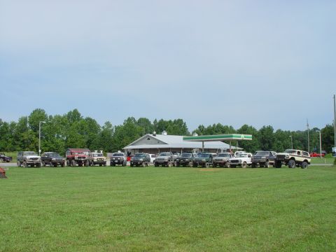 1996 Ford Bronco - A collection of off-road vehicles are gathered in a field near a gas station under a cloudy sky.