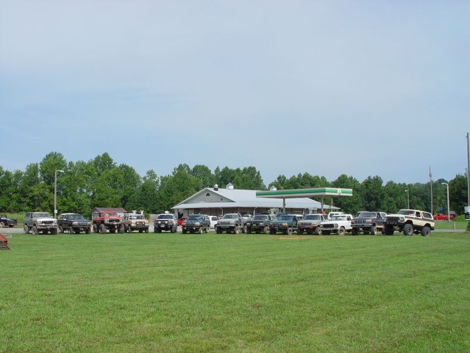 Line of SUVs and trucks parked in a grassy field in front of a gas station