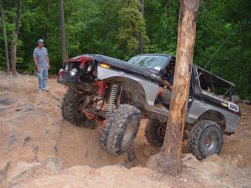 Modified SUV climbing rocky terrain next to a tree. A person stands watching