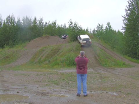 1988 Ford Bronco - A man in a maroon sweatshirt and jeans photographs a white-topped 4x4 descending a steep eroded dirt hill climb, with a second vehicle park…