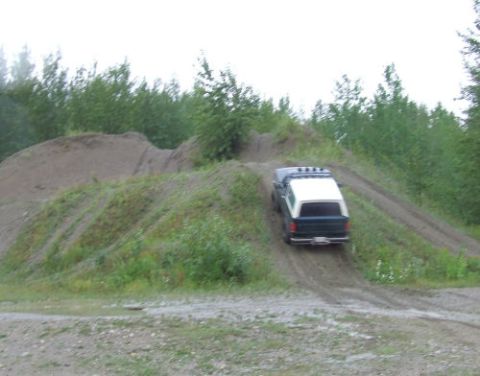 1988 Ford Bronco - A green and white full-size Ford Bronco with a roof light bar descends a steep dirt mound at an off-road play area, seen from behind at the…