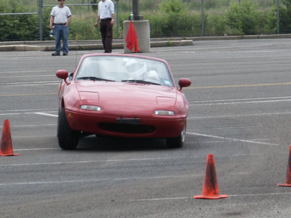 Red Mazda Miata with pop-up headlights driving on a paved course with orange cones