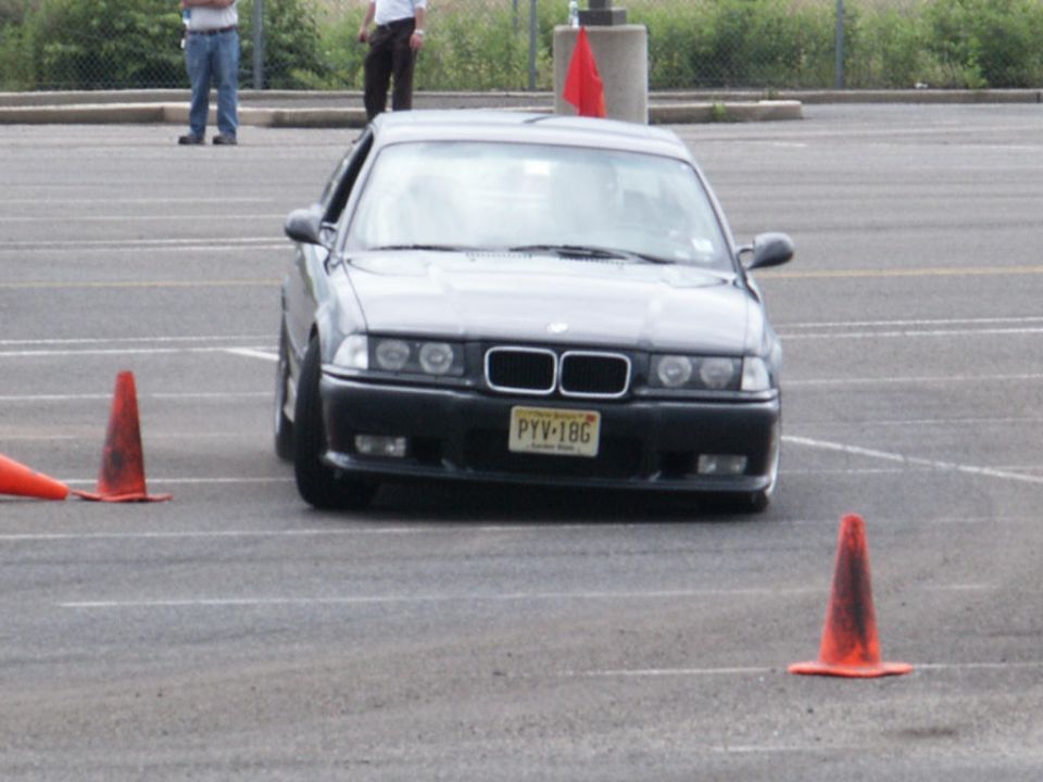 Front view of a dark BMW E36 with a yellow license plate. Orange cones are on the asphalt