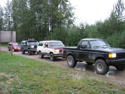 1995 Ford Bronco - A line of SUVs and trucks, including a black Ford Bronco, drive down a muddy dirt road.