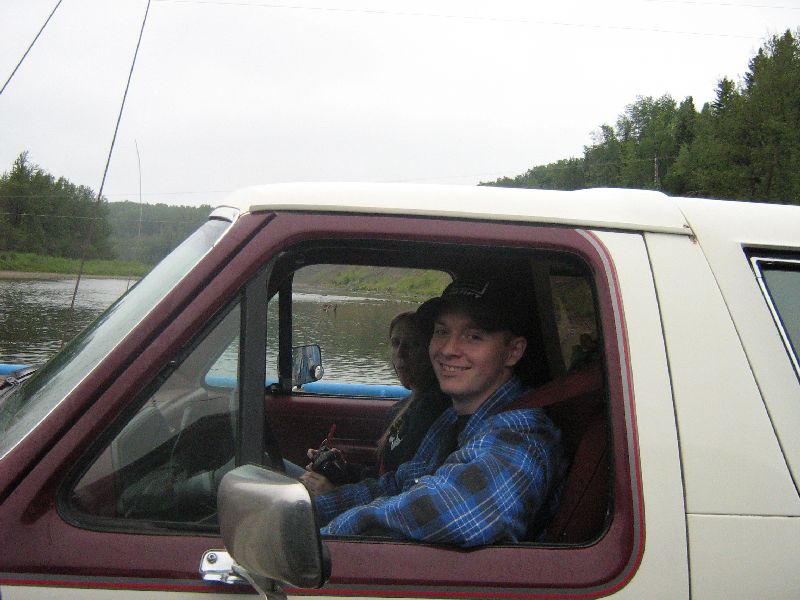 Driver seated in a two-tone maroon and white Ford Bronco parked beside a river